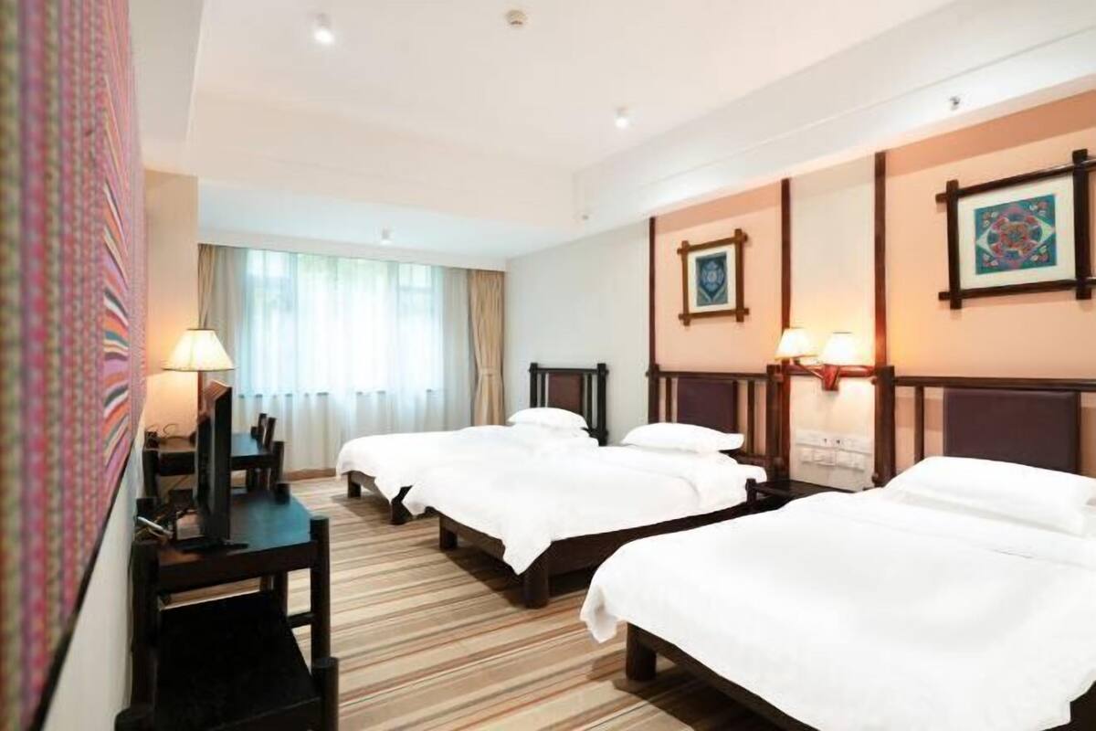 Ein Hotelzimmer mit drei Betten, einem Schreibtisch mit Lampe, einem Fernseher und einem Fenster mit Vorhängen.