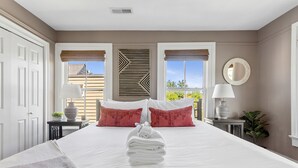 A neatly made bed with white linens, a stack of folded towels, and two red pillows. There is a bedside table with a lamp and a vase.