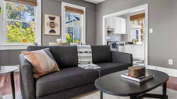 A modern living room with a gray sofa, a coffee table, and a view of the kitchen through an open doorway.
