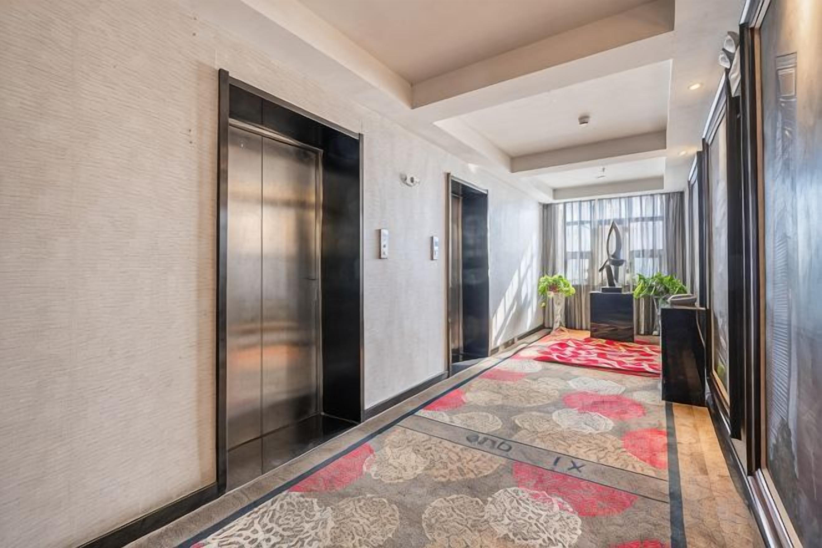 A hotel corridor with elevators, a patterned carpet, and a view into a room with a large window.