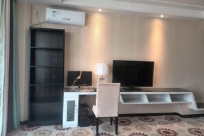 A hotel room with a desk, a chair, a flat-screen TV, a bookshelf, and an air conditioning unit.