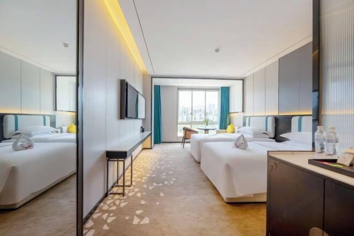 Quarto de hotel com duas camas, uma escrivaninha, uma cadeira e uma janela grande.