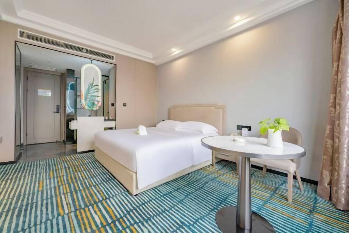 Quarto de hotel com cama, mesa redonda, cadeira, planta em vaso e espelho.