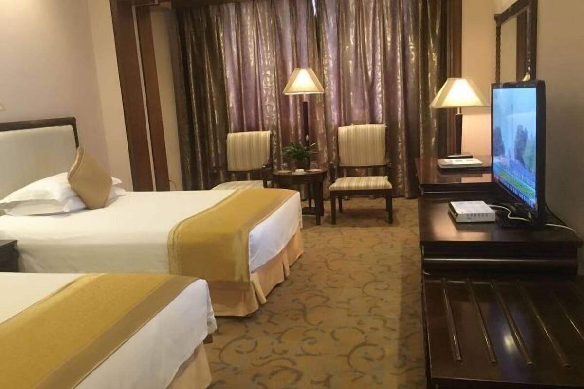 Quarto de hotel com duas camas, televisão, escrivaninha com abajur e uma pequena mesa com planta.