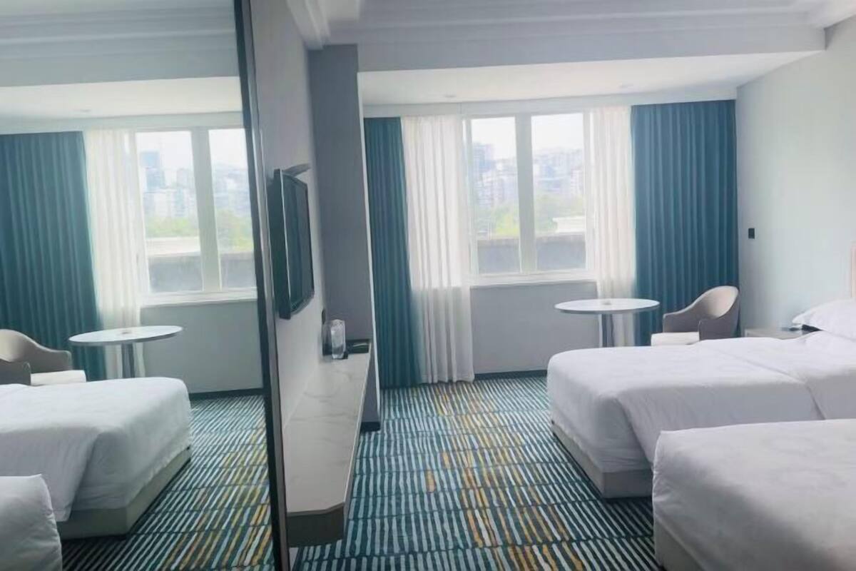 Quarto de hotel com duas camas, uma mesinha, uma cadeira e uma janela grande com cortinas.