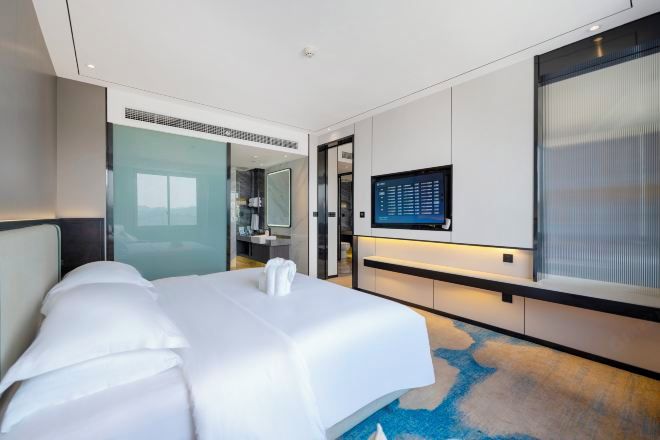 A modern hotel room with a large bed, a flat-screen TV mounted on the wall, and a glass door leading to a bathroom.