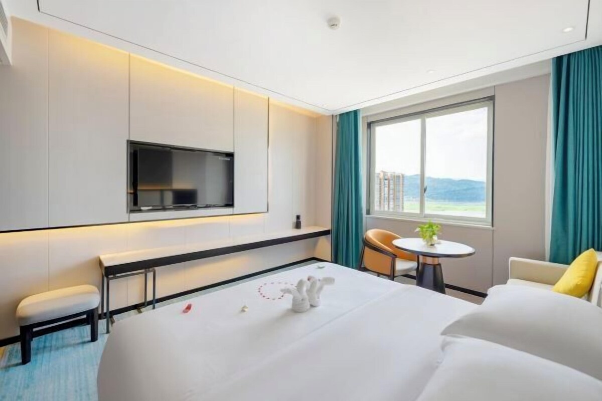 A modern hotel room with a large bed, a TV mounted on the wall, a small table with a plant, and a view of the city through the window.