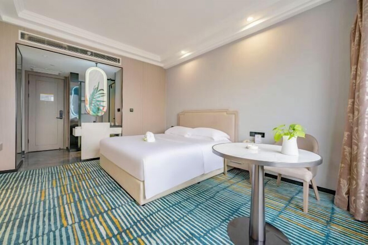 A hotel room with a bed, a round table, a chair, a potted plant, and a mirror.