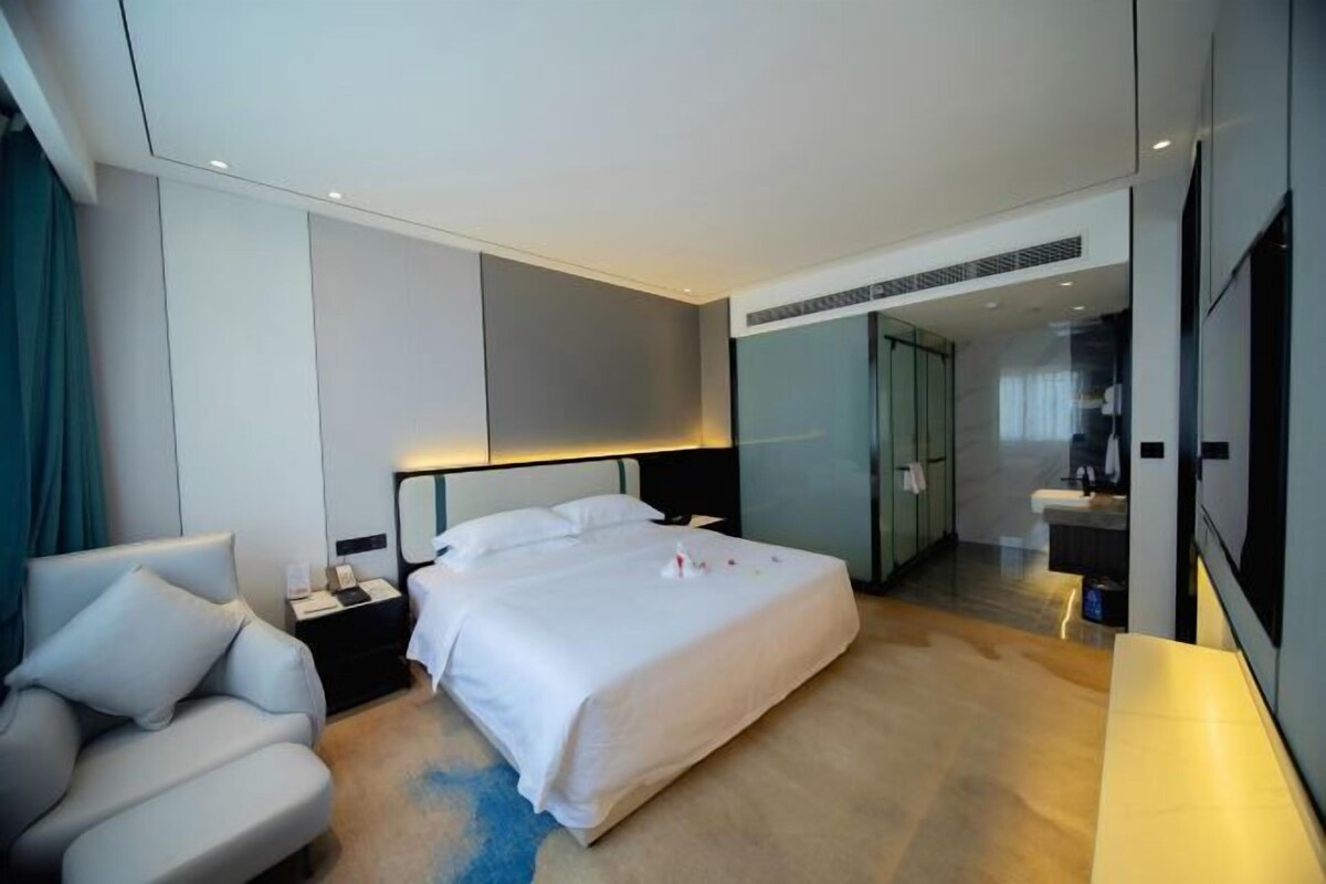 A modern hotel room with a large bed, a bedside table, a seating area, and a bathroom visible through a glass door.