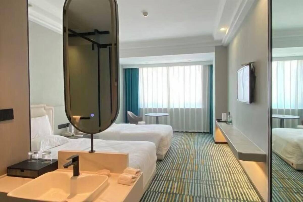 A hotel room with two beds, a large mirror, a sink, a small table, and a window with curtains.