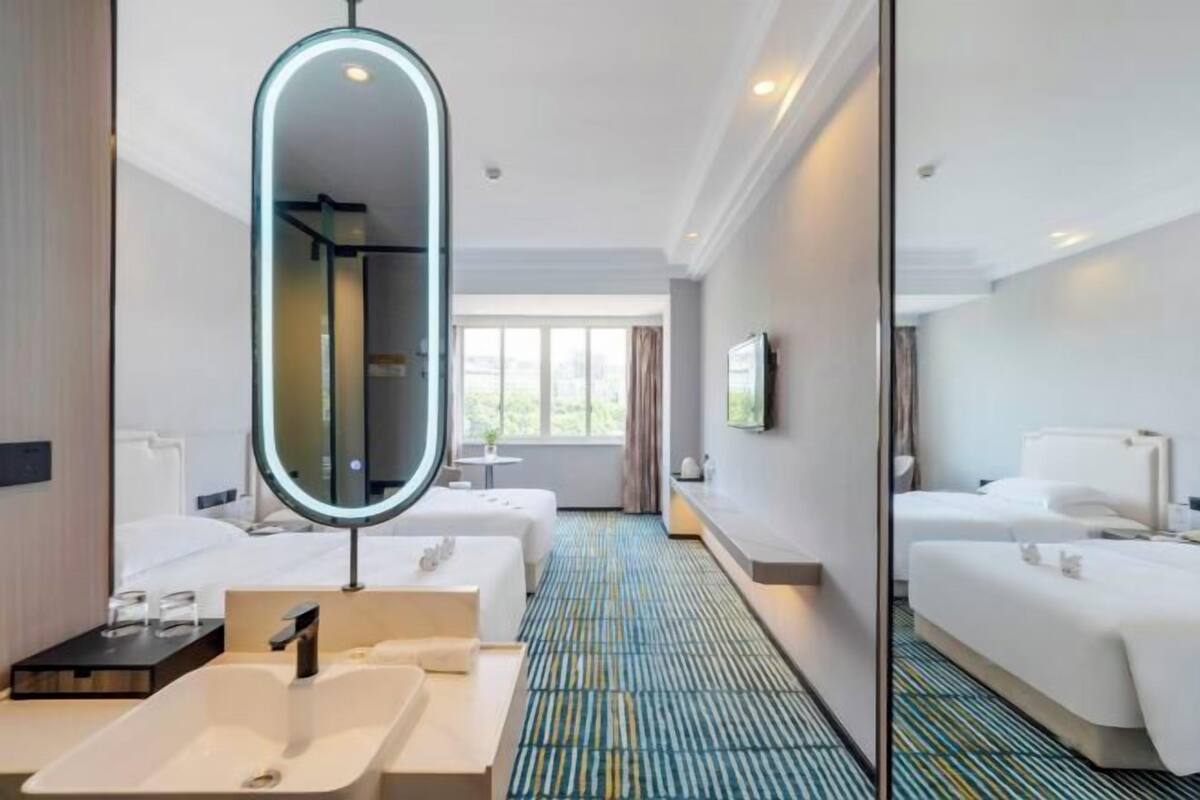 Quarto de hotel com um espelho grande, duas camas, um banheiro com pia e um carpete estampado.