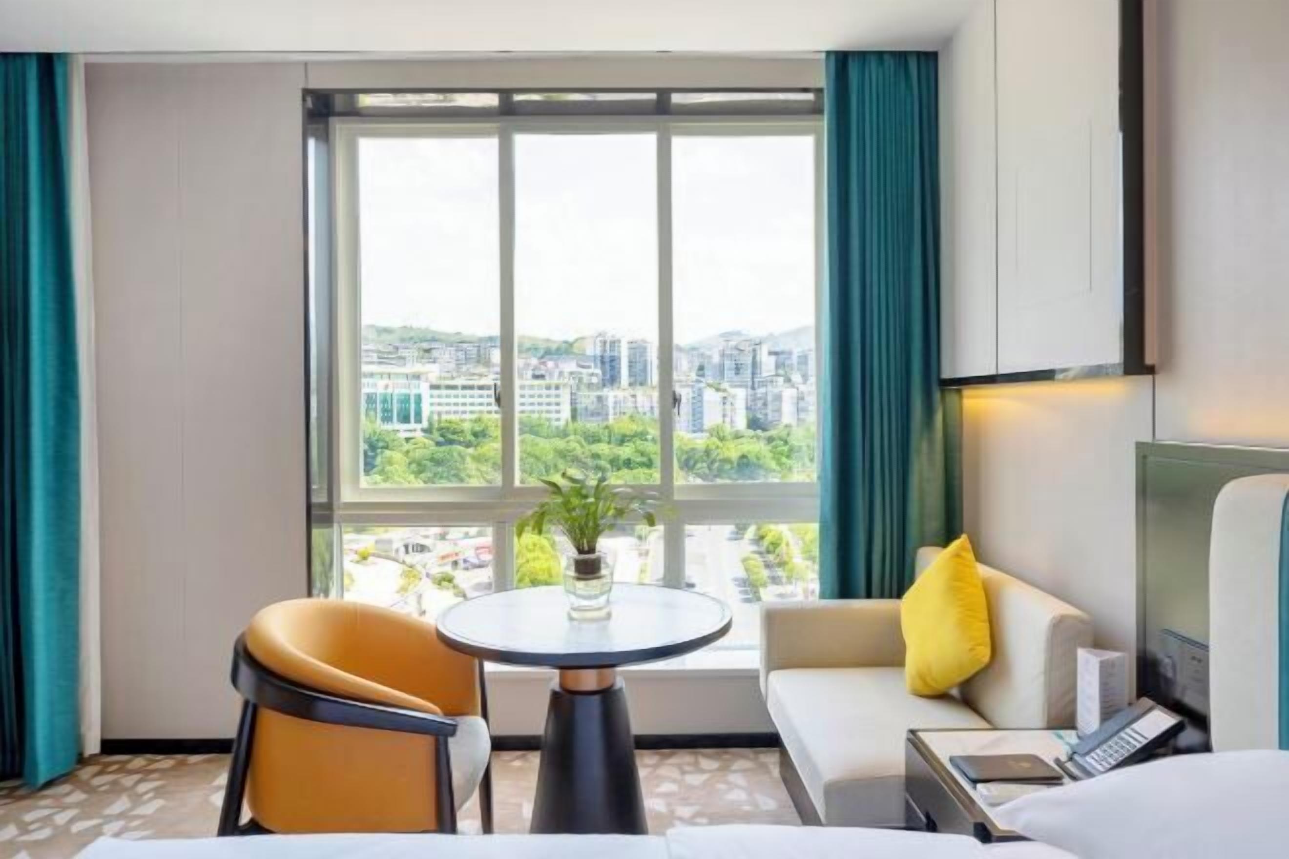 A hotel room with a large window, a round table, a sofa, and a bed.