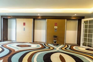Vestíbulo de hotel con alfombra estampada, tres ascensores y un mostrador de recepción.