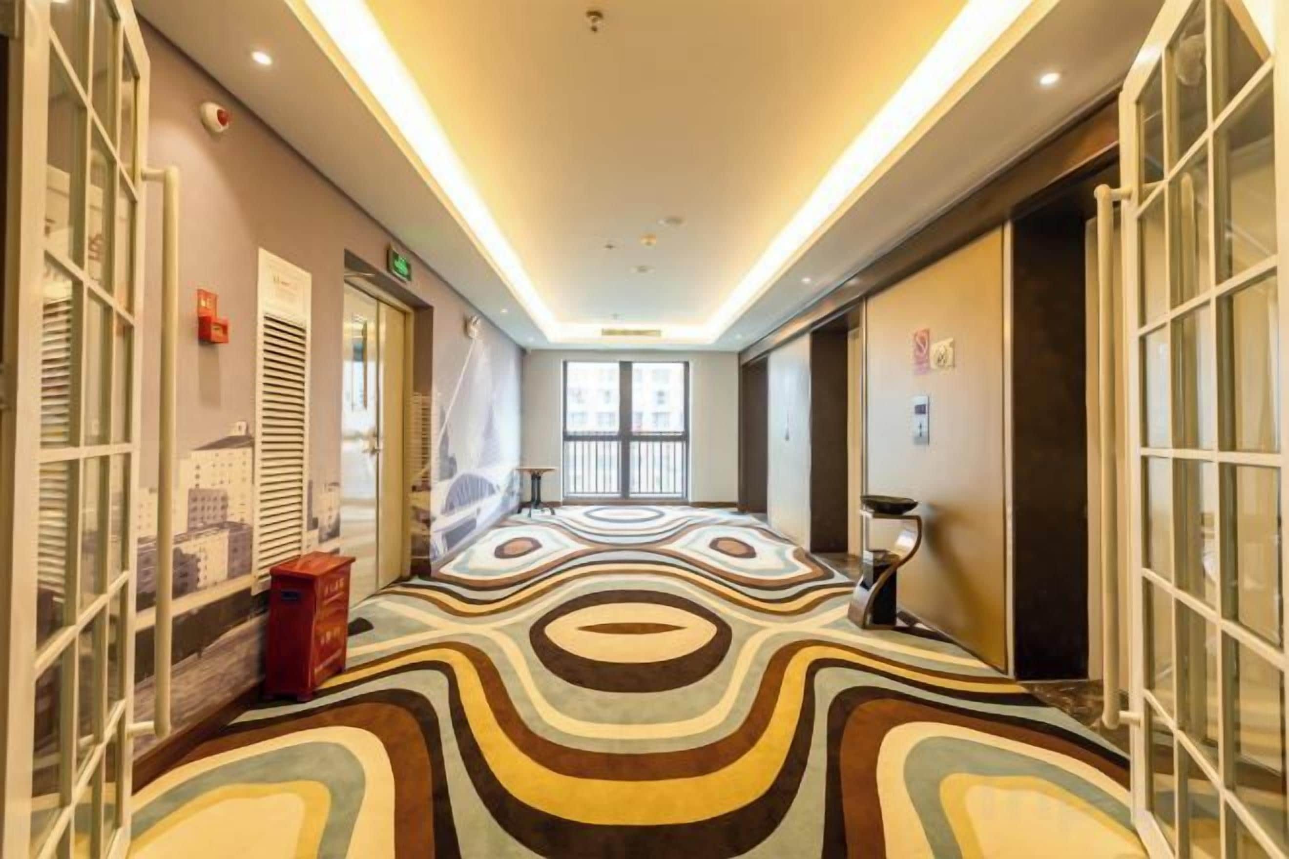 Un couloir d’hôtel avec un tapis à motifs, des portes en verre et une vue sur la ville.