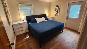 Une chambre à coucher comprenant un lit, des tables de chevet, un ventilateur de plafond, une porte et une fenêtre avec des stores.