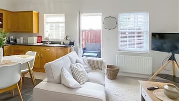 Ein modernes Wohnzimmer mit Sofa, Essbereich mit Tisch und Stühlen, Küche mit Holzausstattung, Fernseher und Fenster mit Jalousien.