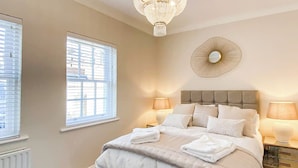 A bedroom with a bed, two bedside lamps, a chandelier, a window with blinds, and a radiator.