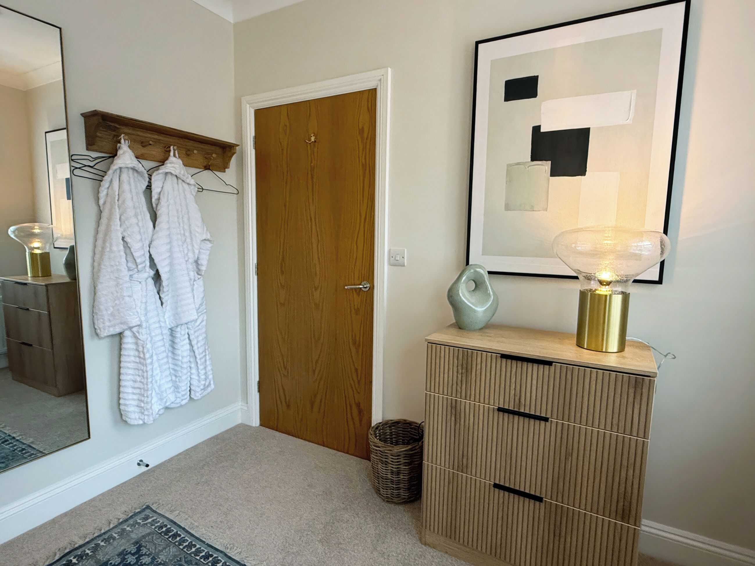 Un couloir avec une porte en bois, un miroir, un porte-manteau blanc, un tableau abstrait encadré, une lampe et un meuble de rangement.