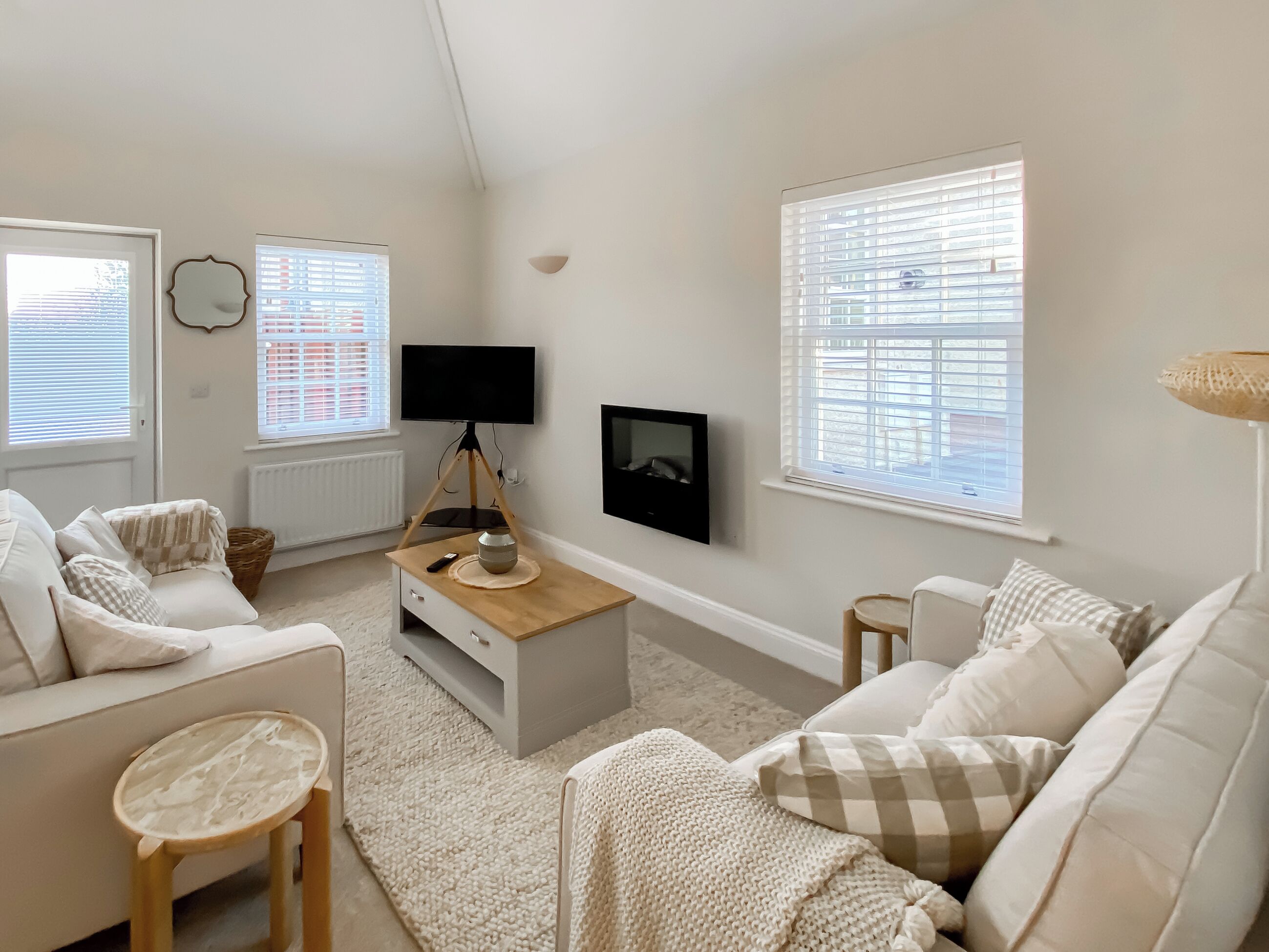 A cozy living room with a white sofa, a coffee table, a TV on a tripod, and large windows with blinds.