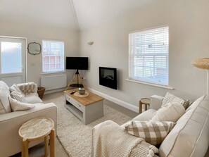 Una sala de estar acogedora con un sofá blanco, una mesa de centro, un televisor sobre un trípode y ventanas grandes con persianas.
