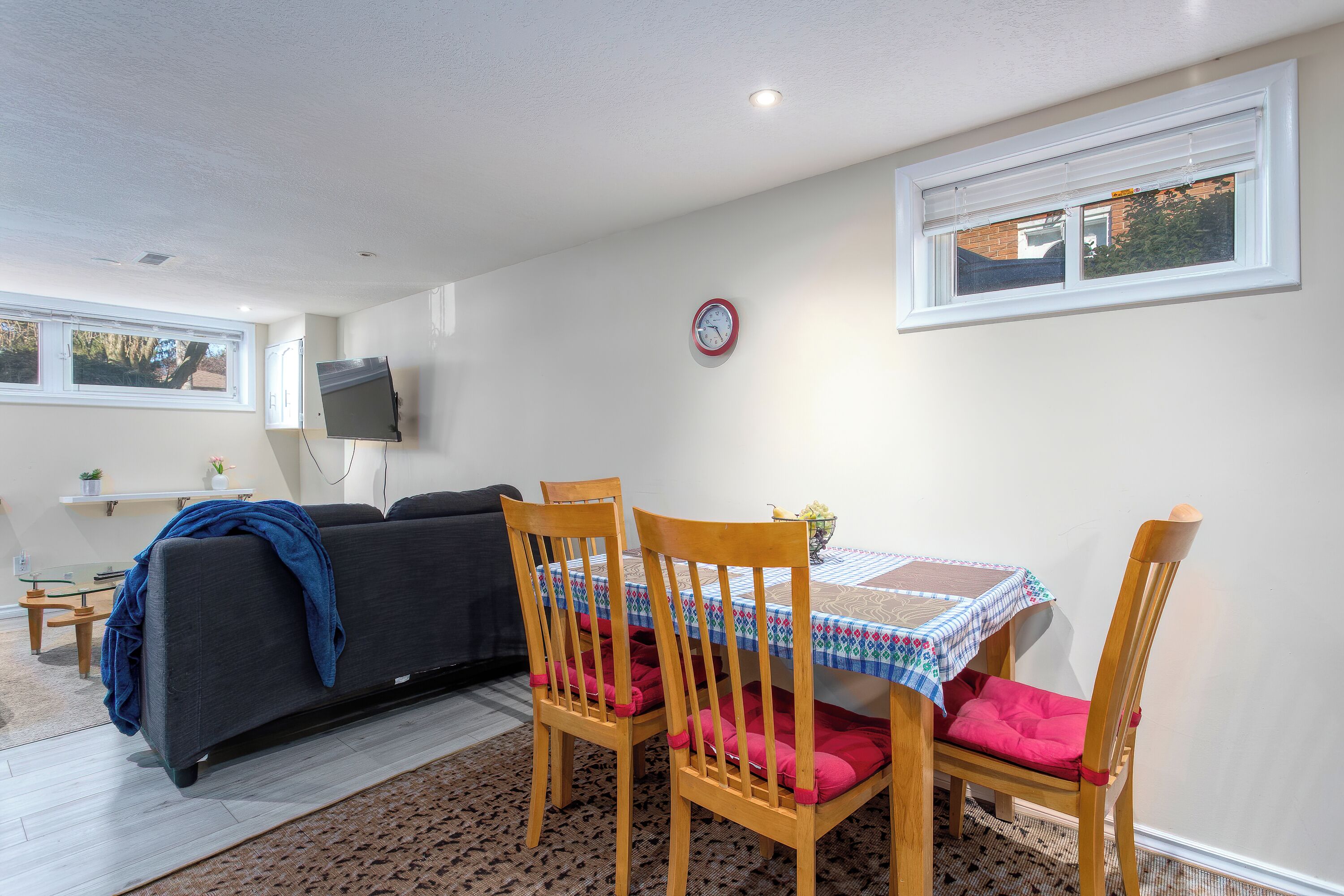 A living room with a sofa, a dining table with chairs, a TV mounted on the wall, and a clock.