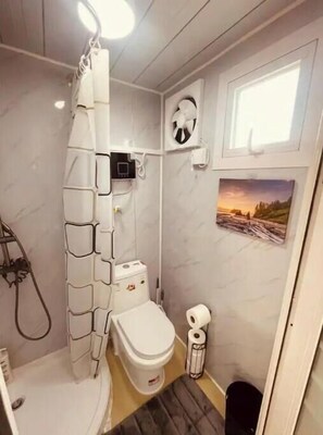 Une petite salle de bain comprenant des toilettes, une douche, une petite fenêtre et un ventilateur fixé au mur.