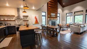 A modern kitchen with a central island, wooden dining table, and bar stools. There is a fireplace, large windows, and a cozy seating area with a sofa and chairs.