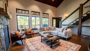A spacious living room with a stone fireplace, large windows, a staircase, and a cozy seating area.