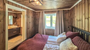 A bedroom with a wooden interior, a bed with a quilted cover, a window with curtains, and a bathroom with a mirror and sink. - Awesome home in Norheimsund with WiFi (Norheimsund)