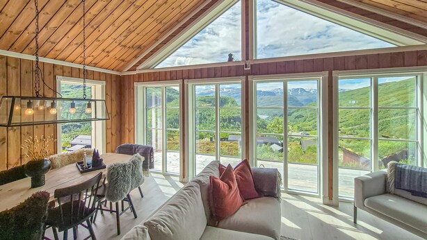 A spacious room with a wooden ceiling, large windows offering a view of green hills and a lake, a dining area with a table and chairs, and a comfortable seating area with a sofa and cushions. - Awesome home in Norheimsund with WiFi (Norheimsund)