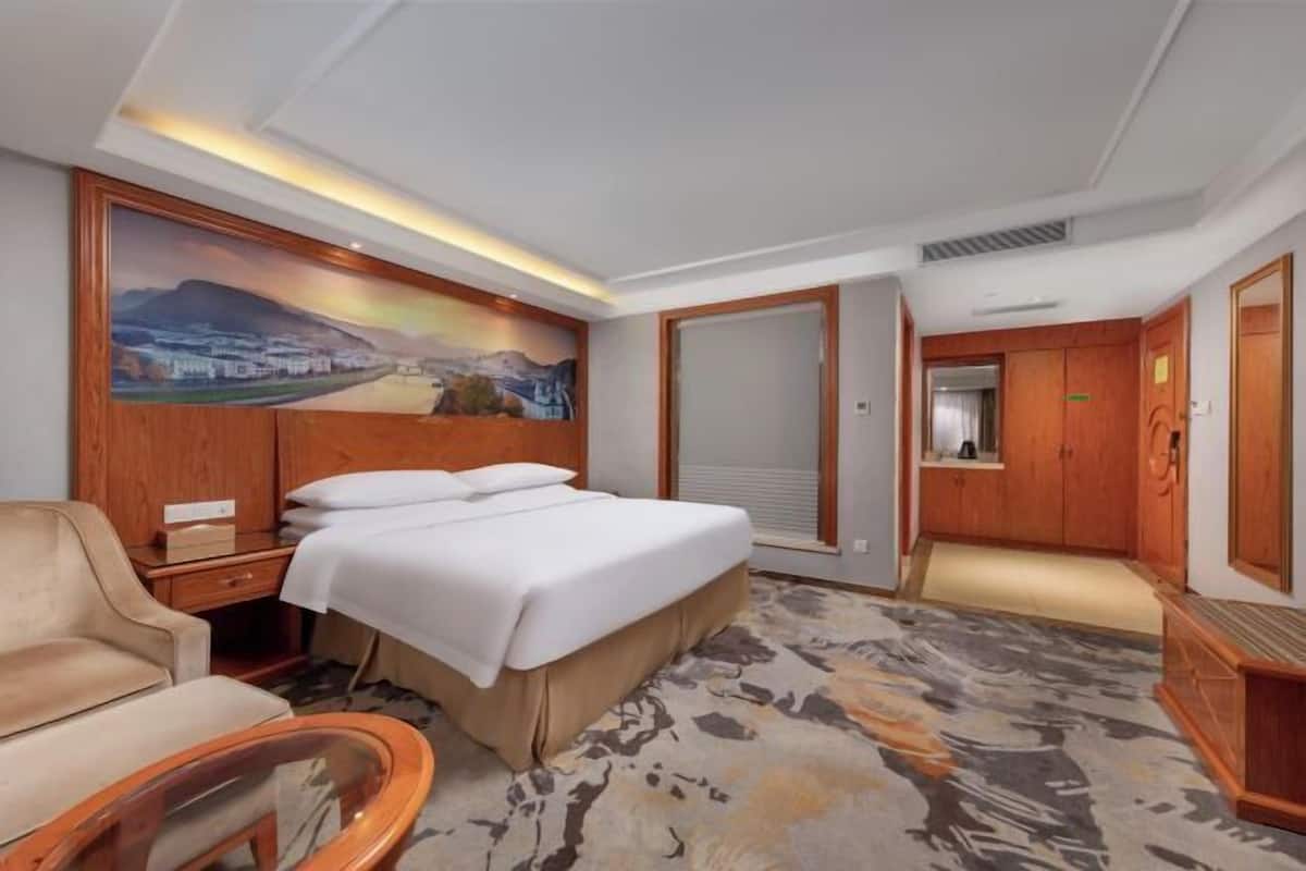 A hotel room with a bed, a desk, a chair, a painting on the wall, and a carpeted floor.