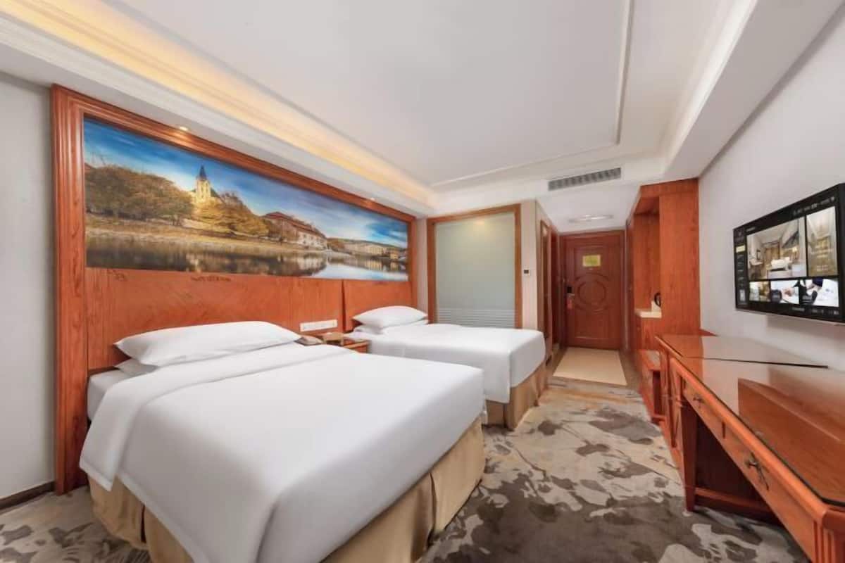 A hotel room with two beds, a painting of a landscape, a television, and a wooden desk.