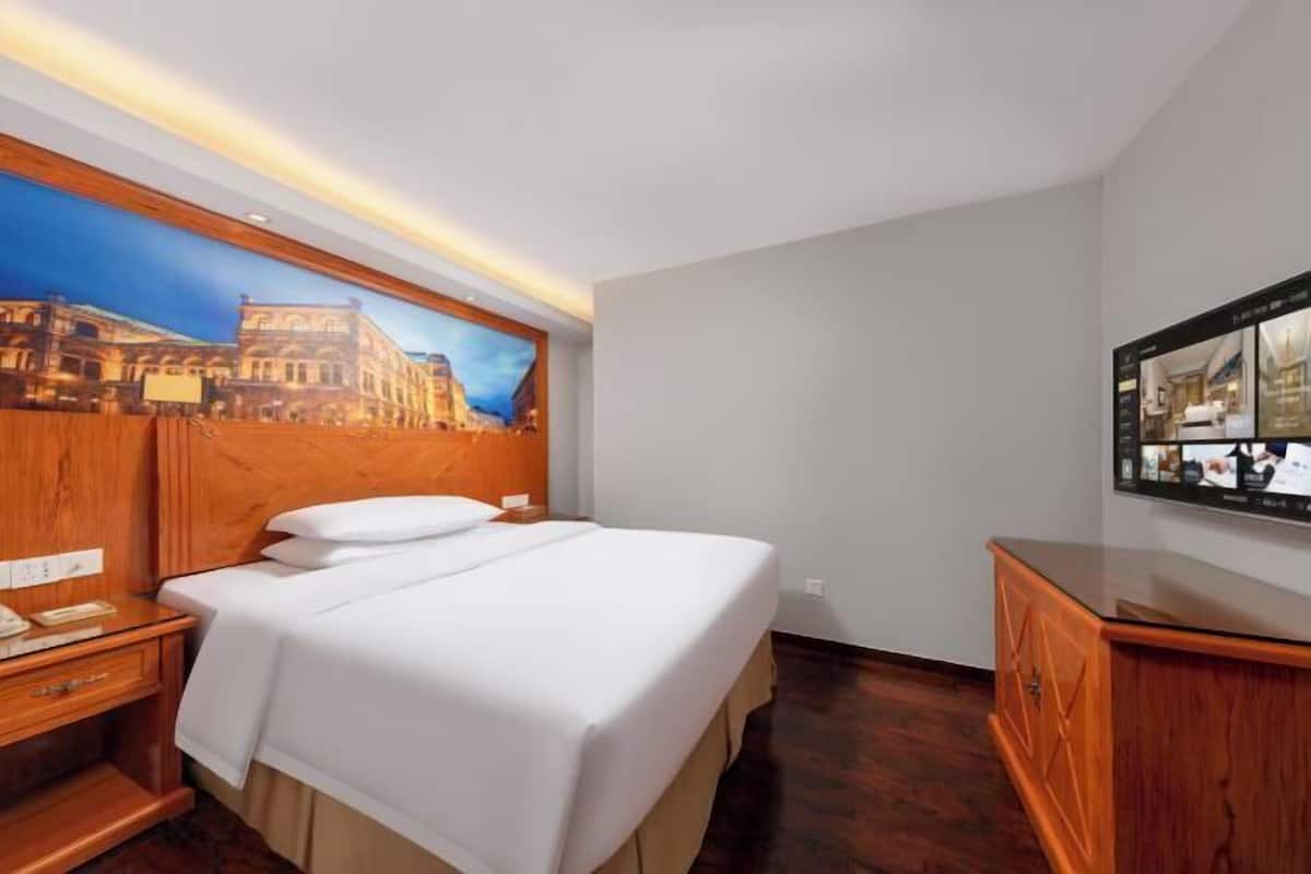 A hotel room with a bed, wooden headboard, a nightstand, a dresser, a wall-mounted TV, and a large window with a cityscape view.