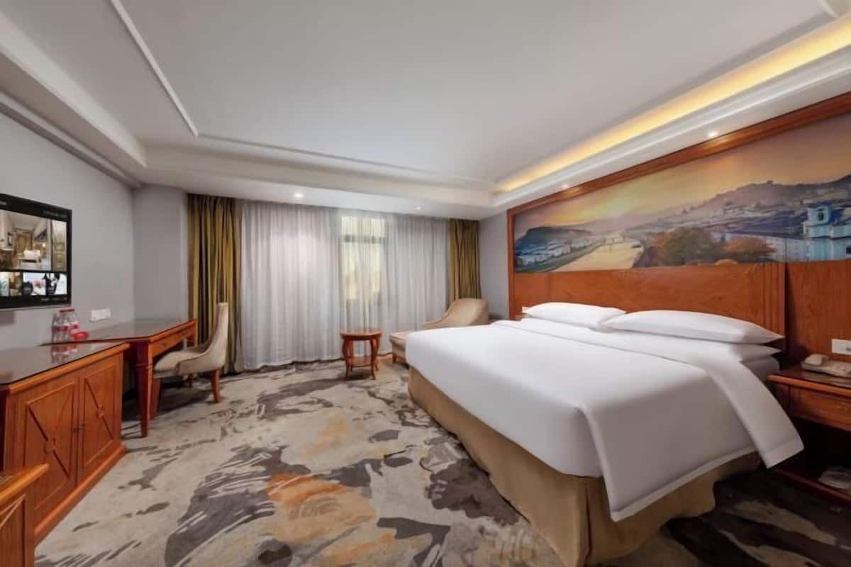 A hotel room with a large bed, a desk, a chair, and a painting on the wall.