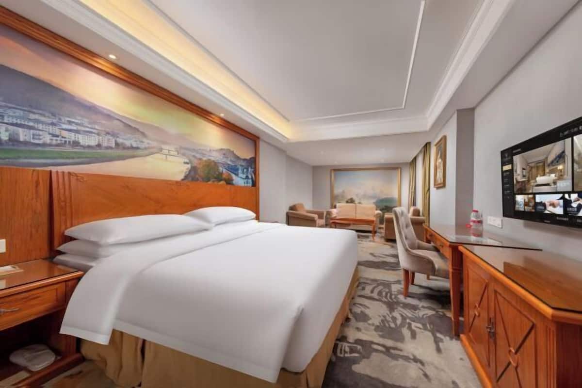 A hotel room with a bed, a desk, a chair, a TV, and a painting on the wall.