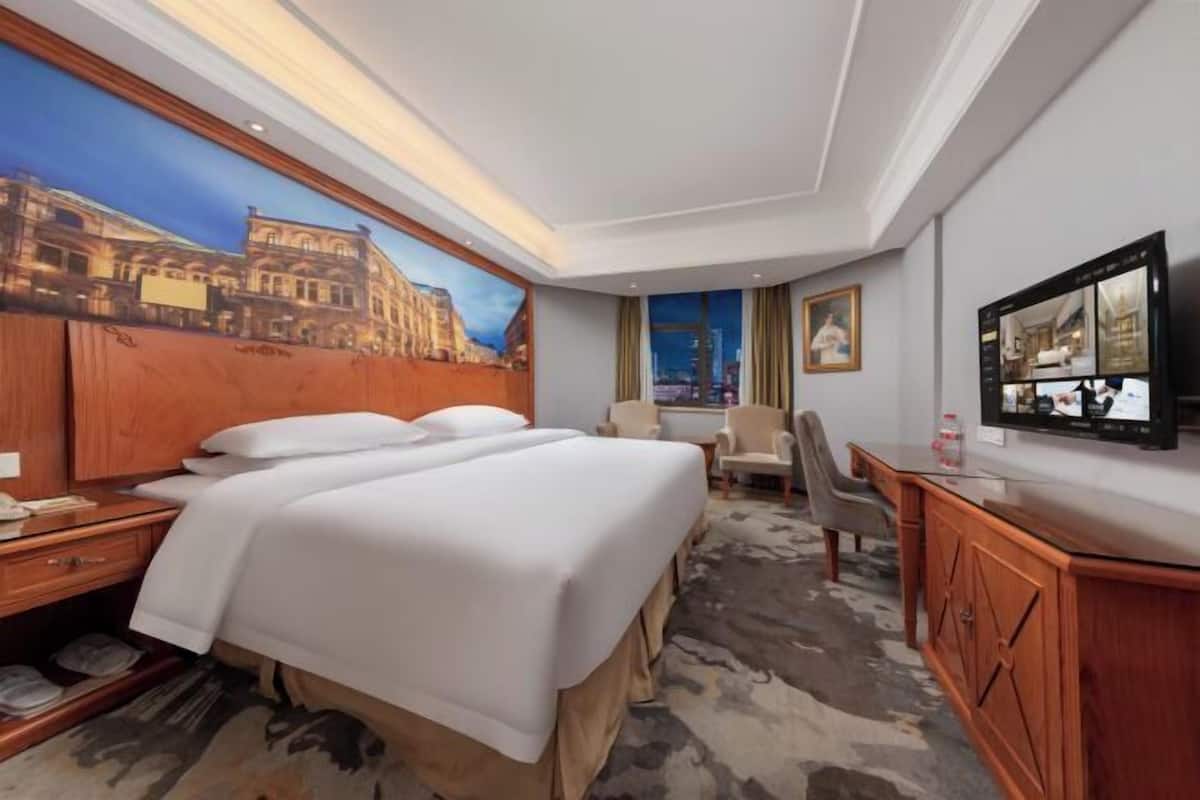 A hotel room with a bed, a desk, a chair, a television, and a large painting on the wall.