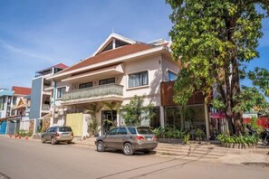 Un immeuble résidentiel de deux étages avec un balcon, des voitures garées et un arbre devant.