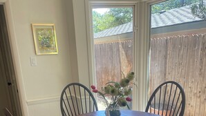 Una mesa de comedor redonda de madera con cuatro sillas negras, un florero con flores y una valla de madera visible a través de la ventana.