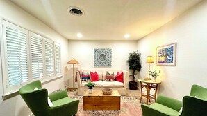 Una sala de estar con un sofá color beige, sillas verdes, una mesa de centro, un tapete estampado, una lámpara de pie, una planta en maceta y obras de arte en las paredes.