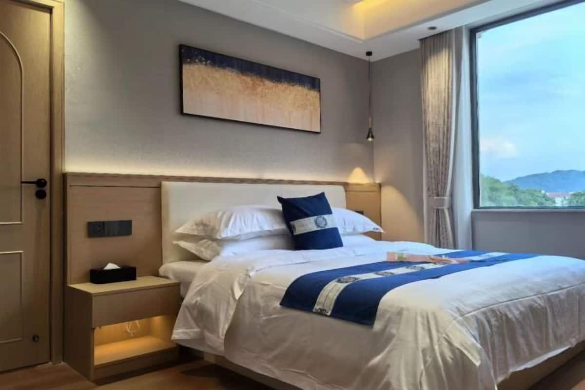 Une chambre d’hôtel avec un grand lit, une table de chevet avec une lampe, un tableau au mur et une fenêtre donnant sur l’extérieur.