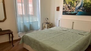 Quarto com cama, mesa de cabeceira, luminária, janela com cortinas leves e pintura de um barco em um penhasco.
