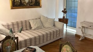 Sala de estar com sofá listrado, um quadro emoldurado acima, uma pequena mesa com um vaso e uma janela com vista para árvores.