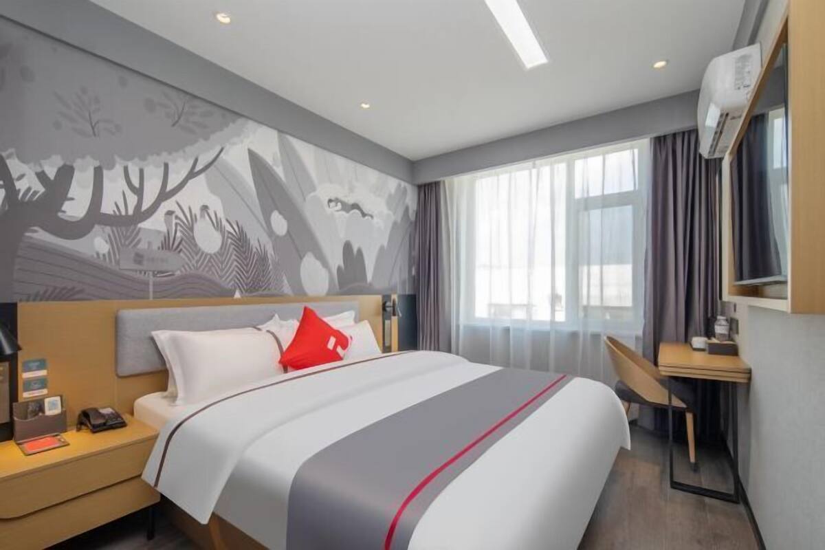 A hotel room with a bed, bedside table, desk, and a large window with curtains.