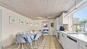 Een moderne keuken met een eethoek, met een lange tafel met stoelen, witte kasten, een raam en een betegelde vloer.
