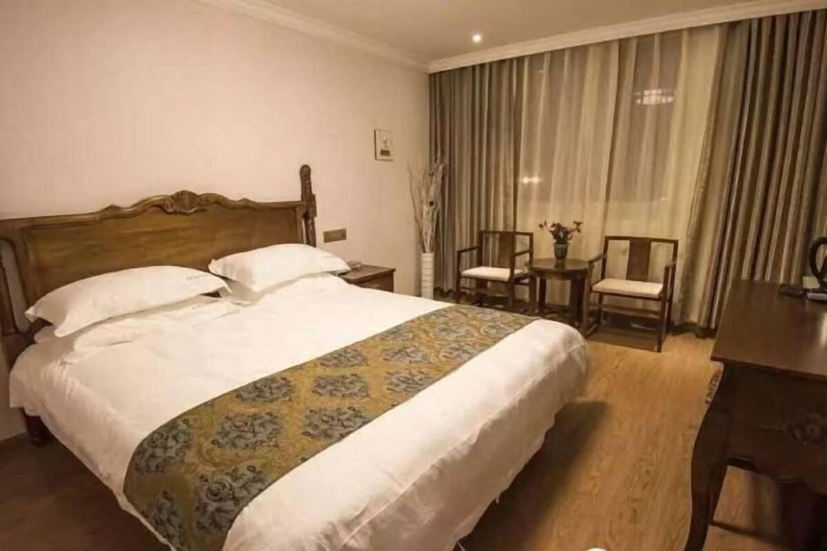 Habitación de hotel con una cama grande, cabecera de madera, una pequeña mesa con sillas y una ventana con cortinas.