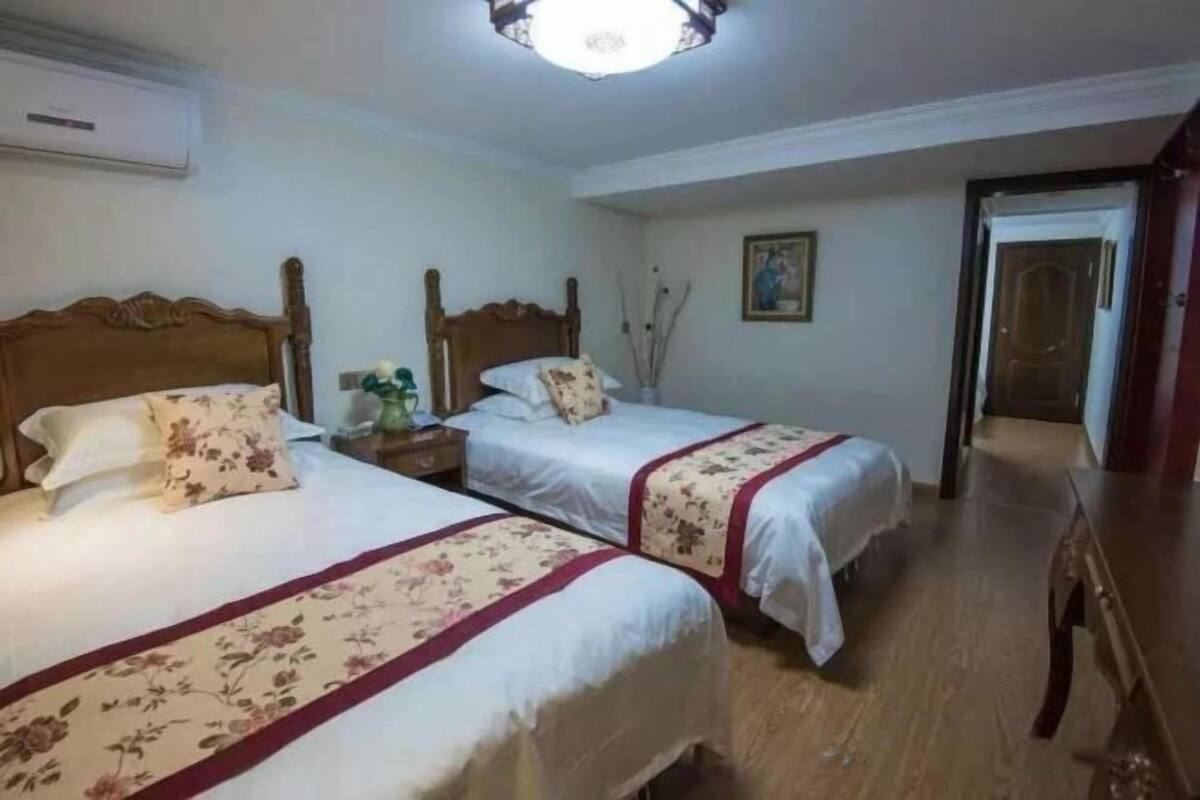 Una habitación con dos camas, cabecero de madera, una mesita de noche, aire acondicionado montado en la pared y una puerta que da a otra habitación.