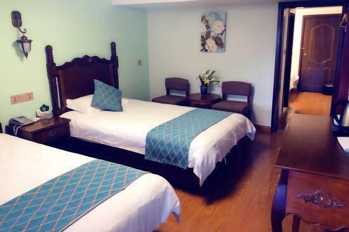 Habitación de hotel con dos camas, cabecero de madera, una mesita con sillas, una lámpara de pared y un cuadro.
