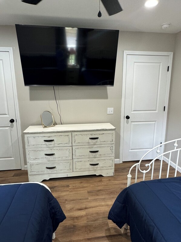 Un dormitorio con una televisión montada en la pared, un aparador blanco con cajones, dos camas con ropa de cama azul y una puerta blanca.