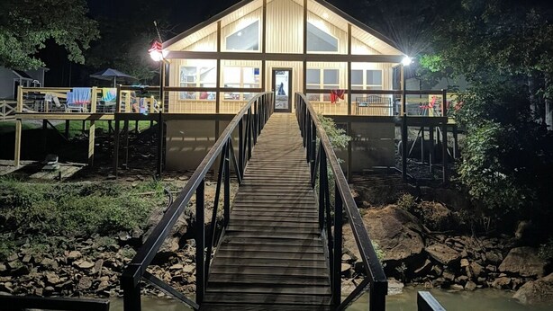 Un puente de madera que conduce a un edificio bien iluminado con grandes ventanales, rodeado de agua y árboles.