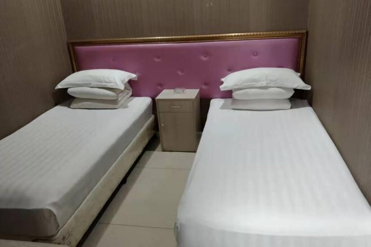Une chambre d’hôtel avec deux lits simples, chacun doté d’un matelas et de coussins blancs, d’une petite table de chevet et d’une tête de lit au motif capitonné.